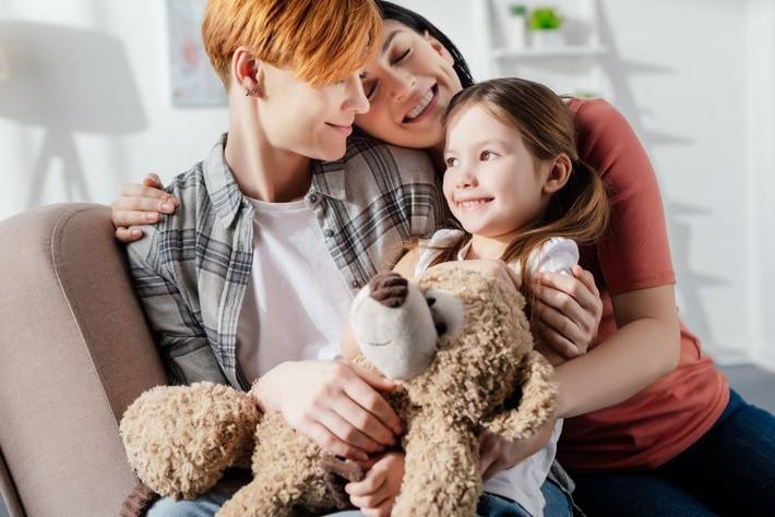 Smiling same sex family embracing daughter with teddy bear on co